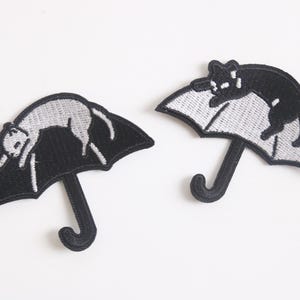 May include: Two black and white embroidered iron-on patches of cats sleeping under umbrellas. The cat on the left is white and the cat on the right is black.