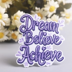 以下が含まれることがあります： 紫色の筆記体で「Dream Believe Achieve」という言葉が刺繍されたパッチ。パッチは白で、紫色のステッチと葉の装飾が施されています。背景にはデイジーがあります。