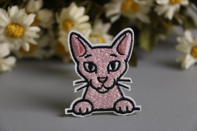以下が含まれることがあります： 黒い縁取りと白い縁取りのある、ピンク色の猫の刺繍パッチ。猫は大きな目、小さな鼻、ひげがあります。背景にはぼやけた白と黄色の花があります。