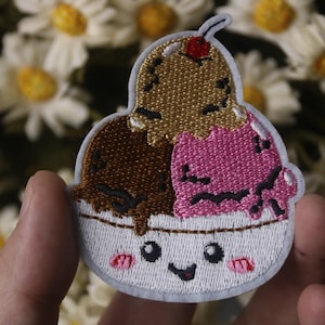 Puede incluir: Parche bordado con un helado sonriente con tres bolas: marrón, dorado y rosa. El helado está en un cuenco blanco con una cara linda y mejillas rosas. Una cereza roja corona la bola dorada.