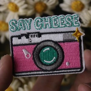 May include: An embroidered patch featuring a pink camera design with the text "SAY CHEESE".