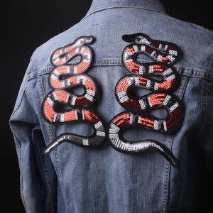 May include: Two red, white and black snake patches with sequin details. The patches are sewn onto a blue denim jacket.