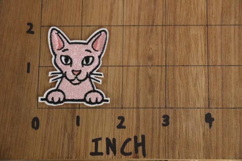 以下が含まれることがあります： 白い縁取りのあるピンク色の猫のパッチ。幅約5cm。猫は笑顔でひげがあり、線から覗いています。パッチはインチのマークが付いた木製の表面にあります。