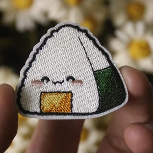 Peut inclure: Un patch brodé blanc et vert en forme de triangle avec un visage souriant et un remplissage jaune. Le patch est une représentation de dessin animé mignon d'une boule de riz japonaise.