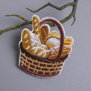 May include: Embroidered patch showing a basket brimming with different breads. The basket is brown with a woven pattern, and the bread is in shades of yellow, white, and brown. A curved branch is in the background.