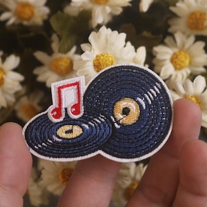 May include: Embroidered patch featuring two vinyl records and a musical note. The records are navy blue with yellow centers and white grooves. The musical note is red and white. The patch is held in front of a background of white flowers.