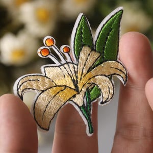 Peut inclure: Une fleur de lys blanche brodée avec des feuilles vertes et des centres orange. La fleur est sur un fond blanc.
