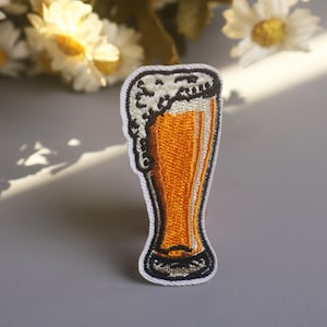Könnte beinhalten: Gestickter Aufnäher eines Bierglases, gefüllt mit bernsteinfarbener Flüssigkeit und bedeckt mit weißem Schaum. Der Aufnäher hat einen weißen Rand und ist wahrscheinlich für Kleidung oder Accessoires bestimmt.