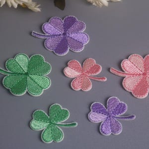 May include: A collection of six embroidered clover patches in various sizes and colors, including two green four-leaf clovers, one pink four-leaf clover, one purple four-leaf clover, one small pink three-leaf clover, and one small purple three-leaf clover.