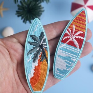 Op de afbeelding: Twee geborduurde surfplatch-patches. E&eacute;n toont een palmboom op een strand met oranje zand en blauw water. De andere heeft een rode en oranje zonsondergang met een palmboom en blauwe golven. Beide zijn lichtblauw omlijnd.