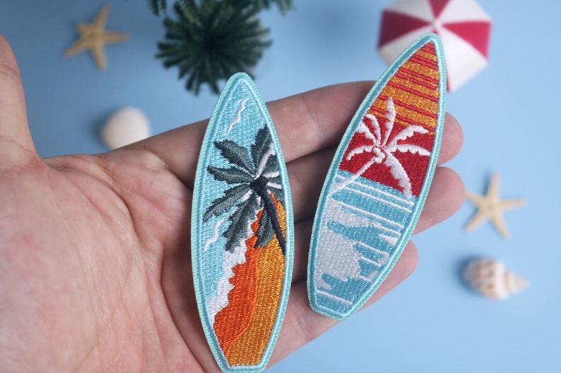 Op de afbeelding: Twee geborduurde surfplatch-patches. E&eacute;n toont een palmboom op een strand met oranje zand en blauw water. De andere heeft een rode en oranje zonsondergang met een palmboom en blauwe golven. Beide zijn lichtblauw omlijnd.
