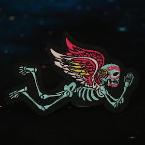 May include: An embroidered patch featuring a skeleton with wings. The skeleton is blue and green, the wings are pink and yellow, and the skull is pink, blue, and yellow.