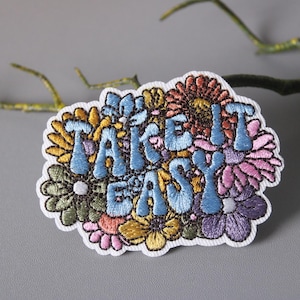 Puede incluir: Parche bordado para planchar con un diseño floral y el texto "Take it Easy" en letras azules.