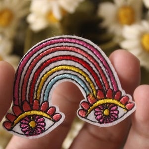 May include: Embroidered rainbow patch with two eyes, each with a pink flower in the center. The rainbow has red, orange, yellow, green, blue, and purple stripes.
