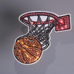 Peut inclure: &Eacute;cusson de basket-ball avec un cerceau et un ballon. Le ballon est marron et le cerceau est rouge et noir, tous deux avec des d&eacute;tails de paillettes. Le filet est noir et blanc.