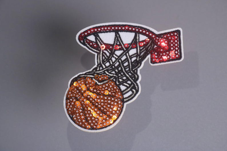 Peut inclure: &Eacute;cusson de basket-ball avec un cerceau et un ballon. Le ballon est marron et le cerceau est rouge et noir, tous deux avec des d&eacute;tails de paillettes. Le filet est noir et blanc.