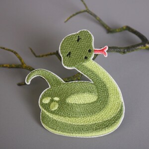 Peut inclure: Patch brodé vert en forme de serpent de dessin animé avec une langue rouge. Le serpent est enroulé en cercle.