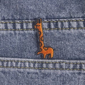 Peut inclure: Un écusson brodé en forme de girafe orange avec un contour marron foncé et des détails de taches circulaires, présentant une texture cousue, fixé sur le tissu en jean bleu d'un pantalon avec des coutures jaunes visibles sur les poches.