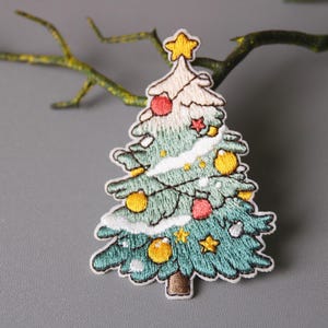 May include: A green and white embroidered Christmas tree patch with yellow and red ornaments, a yellow star on top, and white snow on the branches.