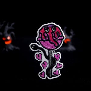 May include: Embroidered patch featuring a whimsical, cartoonish lollipop design. The lollipop head is pink and purple with a face, and the stick is black with pink hearts. The patch has a white border and is set against a black background.