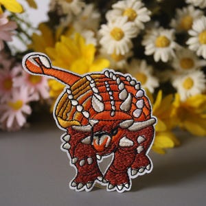 May include: Embroidered iron-on patch of an orange and white ankylosaurus dinosaur. The dinosaur is facing forward with its mouth open and its tail raised.