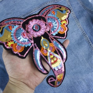 May include: A colorful sequined elephant patch with pink, blue, gold, and orange accents. The patch is on a denim background.