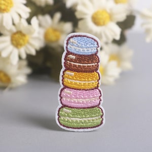 Peut inclure: Écusson brodé représentant une pile de macarons colorés. Le patch présente un design de macaron bleu clair, marron, jaune, rose et vert avec des coutures blanches. En arrière-plan, des marguerites blanches et jaunes.