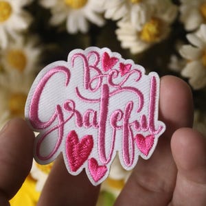 Könnte beinhalten: Gestickter Aufnäher mit den Worten "Be Grateful" in rosa Schrift, akzentuiert mit rosa Herzen. Der Aufnäher ist weiß mit einem rosa Rand. Gänseblümchen sind im Hintergrund.