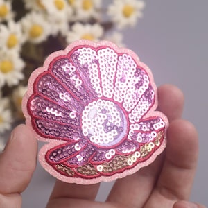 May include: A pink sequined seashell patch with a pearl-like center. The patch has a red outline and gold sequins along the bottom edge. The background is blurred, with white and yellow flowers.