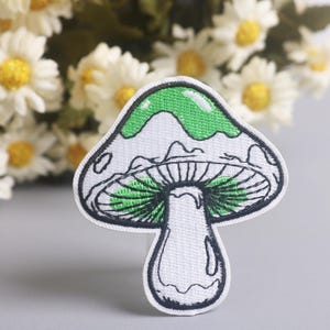 May include: Embroidered mushroom patch with a white background, green top, and black outline. The patch features a detailed design of the mushroom's cap and gills. Daisies are in the background.
