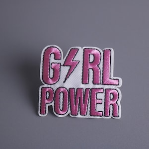 Pode incluir: Remendo de tecido branco com texto bordado em rosa que diz "GIRL POWER" com um raio entre as palavras "GIRL" e "POWER".