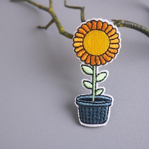 May include: Embroidered iron-on patch of a potted sunflower with a yellow center, orange petals, green leaves, and a dark blue pot.