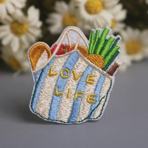 May include: Embroidered patch featuring a blue and white striped tote bag filled with food items. The bag contains a loaf of bread, a tomato, pineapple, and green vegetables. The words "LOVE LIFE" are embroidered in gold.