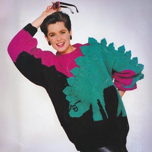 May include: A colorful knitted sweater featuring a dinosaur design. The sweater has a black, pink, and teal color scheme. The dinosaur design is prominently displayed on the front and sleeves. The sweater has a round neckline and long sleeves.