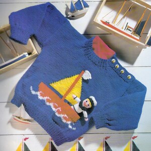 May include: Blue knitted sweater with a nautical theme. The sweater features a sailboat with a yellow sail and a sailor graphic. The collar is red, and the shoulder has four buttons. Decorative wooden sailboats are in the background.