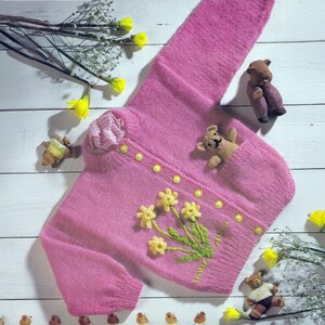 May include: A pink knitted baby cardigan with yellow flower and green stem embroidery. The cardigan has a pocket with a small teddy bear inside. The cardigan has yellow buttons and a decorative flower on the shoulder.