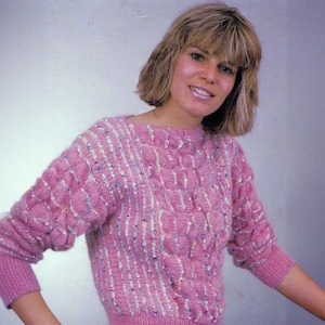 May include: A pink knitted sweater with a textured pattern. The sweater features a round neckline and long sleeves with ribbed cuffs. The knit design includes vertical stripes and raised, square-like sections, with colorful flecks throughout.