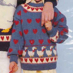 Può includere: Un maglione blu lavorato a maglia con cuori rossi e disegni di oche bianche. Il maglione ha un colletto e polsini a coste con motivi rossi e bianchi. Il maglione è probabilmente per un bambino.