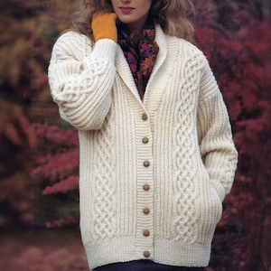 May include: Cream cable-knit cardigan with a shawl collar and wooden buttons. The sweater has intricate cable patterns on the sleeves and front panels. The model is wearing a patterned scarf and yellow gloves.
