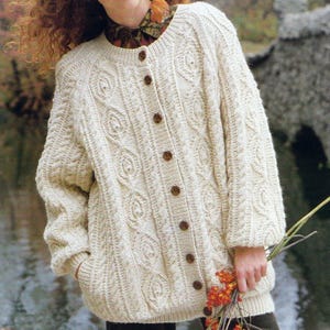 May include: Cream-colored, button-down cardigan sweater with a cable-knit design. The sweater has a round neckline, long sleeves, and brown buttons. The person is holding a small bouquet of orange flowers.