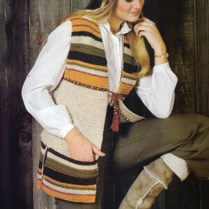 May include: A crochet vest with horizontal stripes in shades of orange, green, and cream. The vest has a cream-colored body with pockets and a tie closure. The model is wearing a white shirt and olive-green pants.