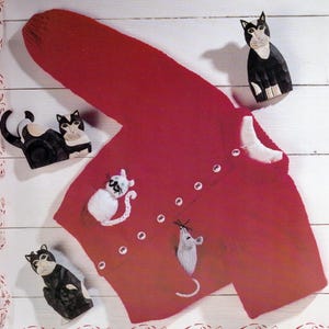 May include: A red knitted cardigan with white button closures and white cat and mouse appliqués. The cardigan is surrounded by carved wooden cat figurines in black and white.