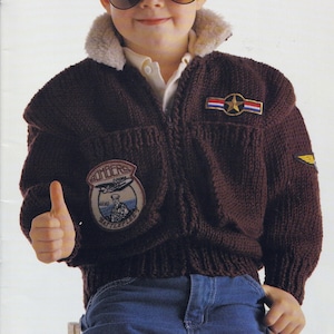 Könnte beinhalten: Gestrickte braune Bomberjacke mit cremefarbenem Kunstfellkragen. Die Jacke hat Aufnäher mit militärischen Motiven und dem Wort "Bombers". Die Jacke wird mit einer blauen Jeans getragen.