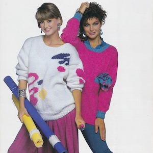 May include: Two women in 1980s fashion. One wears a white sweater with pink, blue, and yellow abstract shapes. The other wears a bright pink sweater with blue trim. One woman holds rolled-up paper.