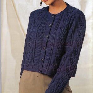 May include: A navy blue cardigan with a round neck and button closure, featuring detailed cable knit and textured panels. The long-sleeved sweater is cropped at the waist. The garment is paired with a tan skirt or trousers. The sweater is made of a knitted fabric.
