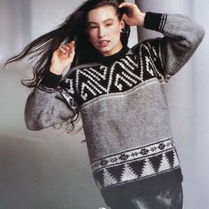 May include: A grey jumper featuring black and white geometric patterns across the chest and lower hem. The jumper has a black collar and cuffs. The model is wearing the jumper and has long dark hair.