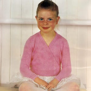 May include: A young person wearing a pink wrap-around cardigan and a white tutu skirt. The cardigan has a V-neck and long sleeves. The person is sitting in front of a white background.