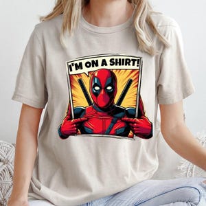 May include: A beige t-shirt featuring a graphic print of Deadpool in a comic book style frame. The text "I'M ON A SHIRT!" is above Deadpool.