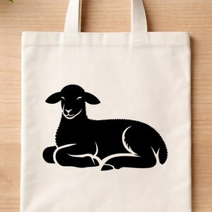 May include: A cream-colored tote bag featuring a black silhouette of a lamb in a resting position. The lamb has detailed fur and is lying down. The bag has two cream-colored handles.