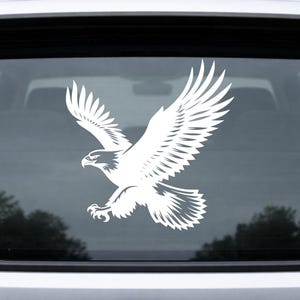 May include: White vinyl decal of an eagle in flight, with outstretched wings and detailed feathering. The eagle is depicted in a dynamic pose, suitable for application on a car window or other smooth surfaces. The design is a silhouette.
